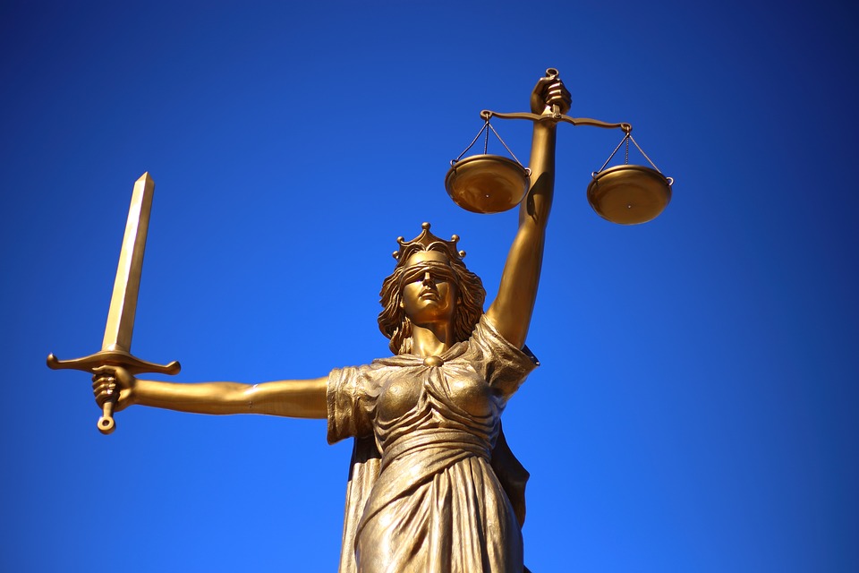A golden statue of Lady Justice holding a sword in her right hand and balanced scales in her left hand. She wears a blindfold and a toga, set against a clear blue sky.