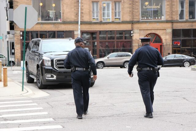 Two police officers in uniform walking down a city street. They are near parked vehicles, including an SUV, and in front of a building with large windows, one of which has an "OPEN" sign. The road features a crosswalk and street signs.