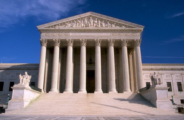 The image shows the front of the United States Supreme Court building. It features a classical design with eight large columns and a triangular pediment. Steps lead up to the entrance, and sculptures are positioned on either side.