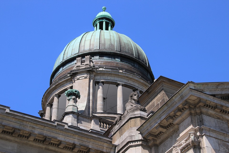 Historic building with a large green dome and classical architectural details, including stone statues and columns, set against a clear blue sky.