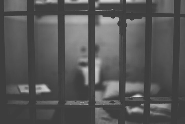 Black and white photo of a prison cell viewed through metal bars. Inside, there's a bed with striped bedding, a toilet, and a small shelf with books. The dim lighting and stark setting create a somber atmosphere.