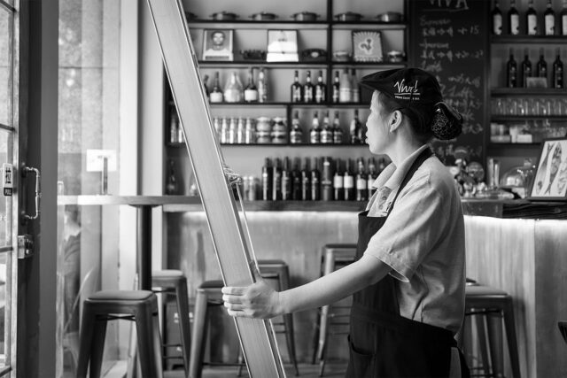 A woman wearing an apron and cap stands inside a café, holding a large frame. She looks towards the open glass door. Shelves filled with bottles and jars cover the wall behind her. The setting appears calm and organized.