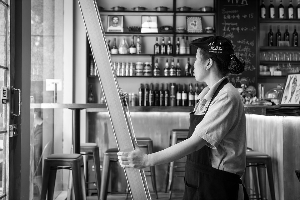 A woman wearing an apron and cap stands inside a café, holding a large frame. She looks towards the open glass door. Shelves filled with bottles and jars cover the wall behind her. The setting appears calm and organized.