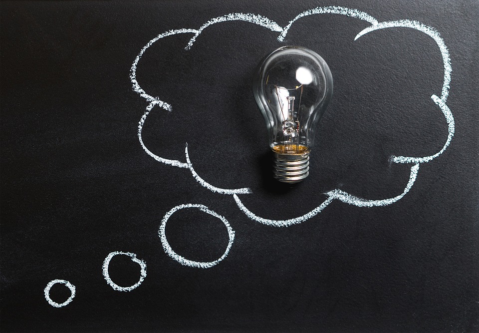 A clear lightbulb is placed on a blackboard with a white chalk drawing of a thought bubble extending from it, symbolizing an idea or a moment of inspiration.