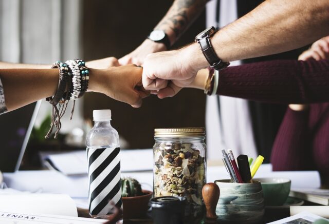 Four people fist-bumping over a table cluttered with items such as a water bottle, a jar of snacks, papers, and other small objects. They wear various bracelets and watches. The scene suggests teamwork or camaraderie.