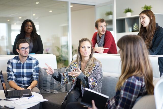 A group of six people are engaged in a discussion in an office setting. One woman is speaking animatedly, gesturing with her hands. Others are seated or standing around, listening attentively. Laptops and notebooks are visible.