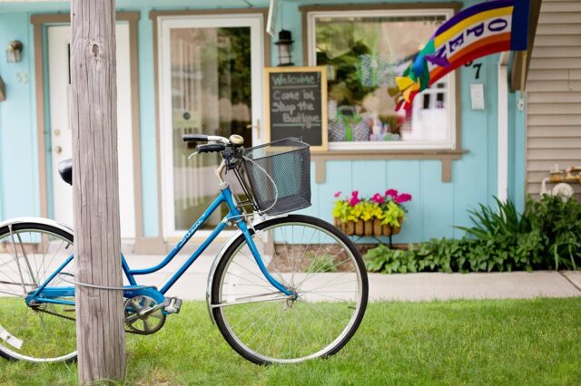 A blue bicycle with a front basket is parked on a grassy area near a pole. Behind it, a small boutique with a sign inviting people to come in and shop displays a colorful flag and flower boxes in front of a light blue storefront.