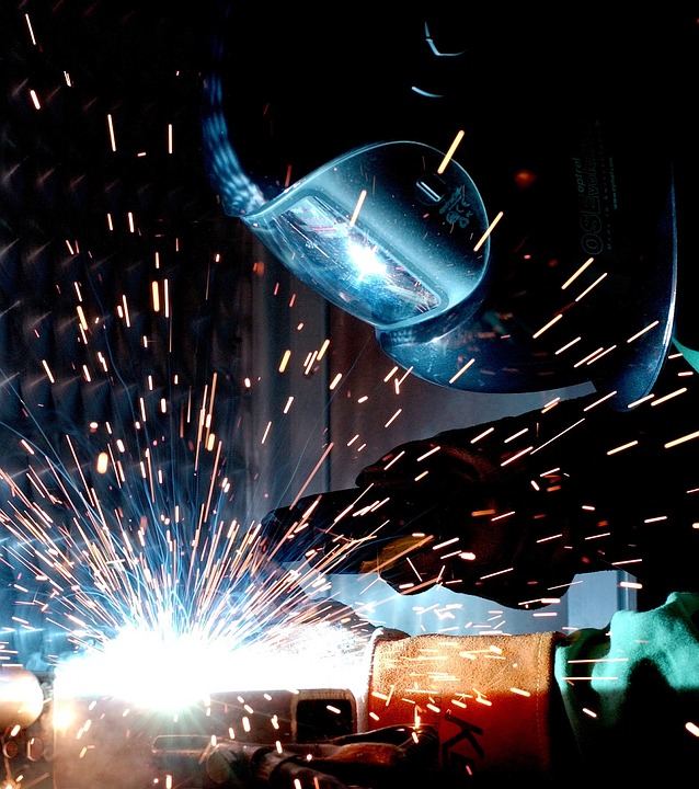 A person wearing protective gear and a welding helmet is performing welding, with bright sparks flying from the welding torch. The image shows intense blue and orange light illuminating the scene.