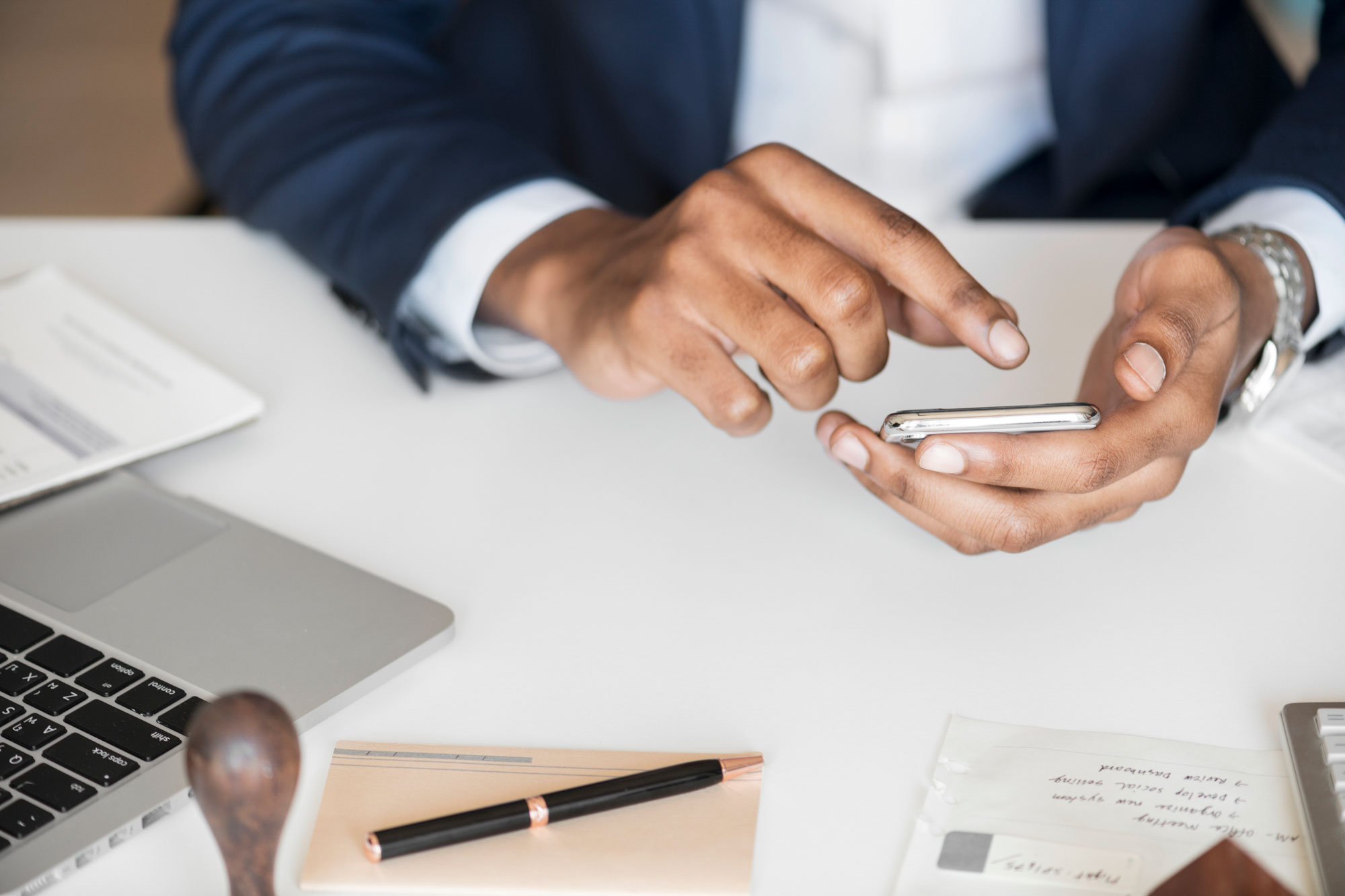 A person wearing a dark suit uses a smartphone at a desk, likely an attorney specializing in business law. Nearby are a laptop, a pen, papers, and a sealed envelope, all contributing to the professional work environment.