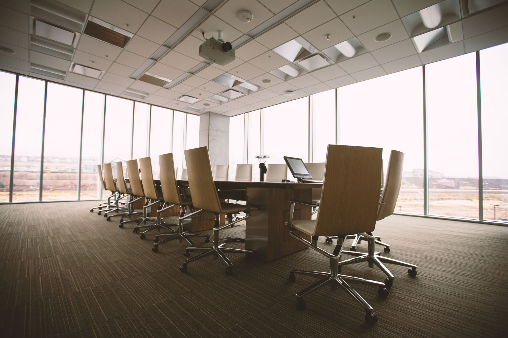 A large, empty conference room with a long wooden table surrounded by swivel chairs awaits a business valuations attorney. The room features floor-to-ceiling windows that offer a stunning view of the exterior landscape, bathing the space in bright natural light.