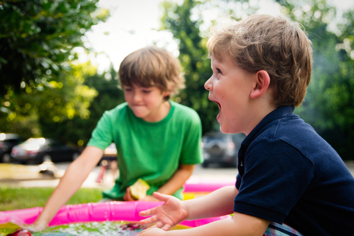 Two young boys play outdoors near a small inflatable pool. One boy in a navy shirt is smiling and gesturing excitedly, while the other, in a green shirt, focuses on the pool. As they enjoy their day under the watchful eye of their child support attorney parent, trees and cars blur in the background.