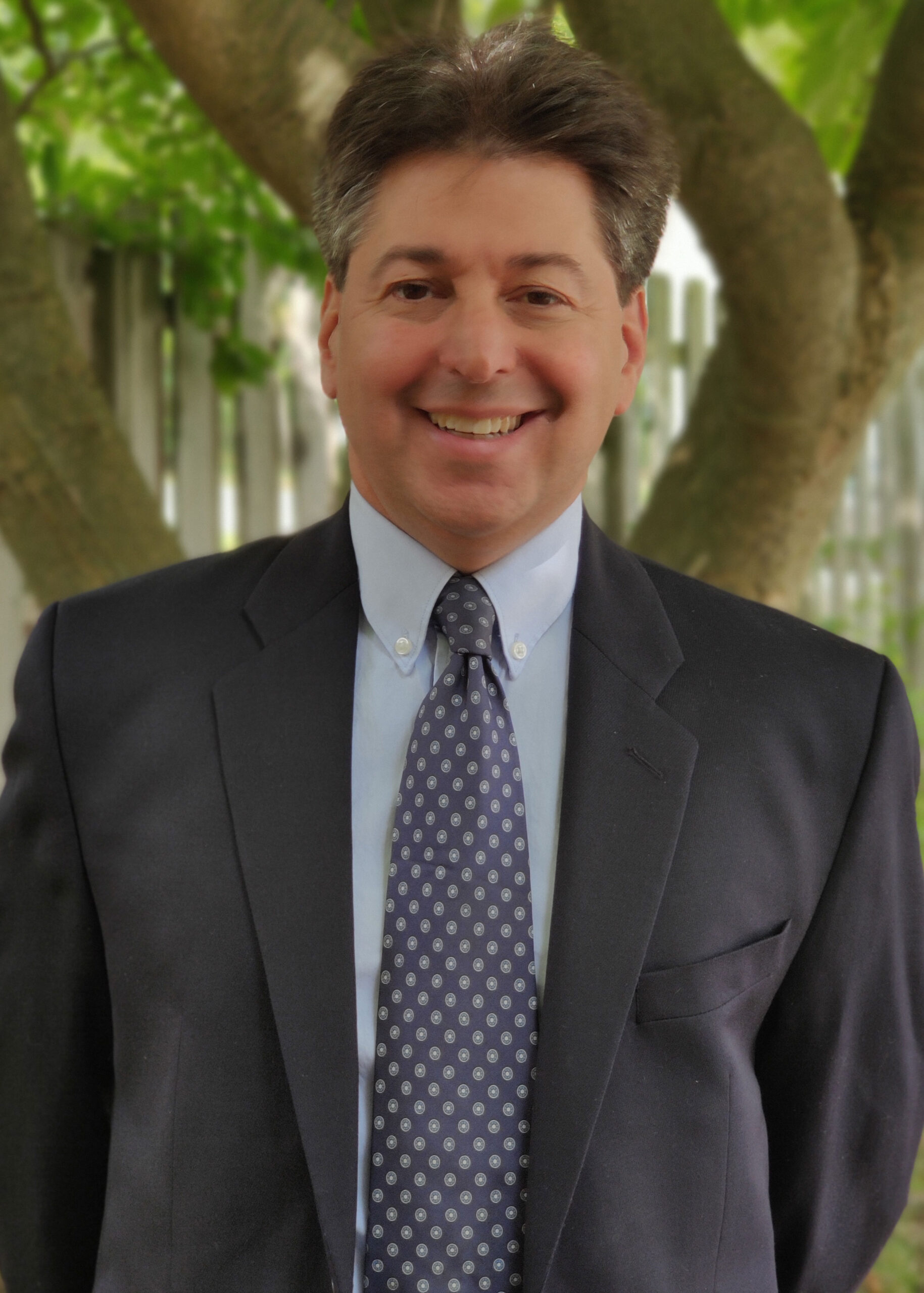 A man in a dark suit, light blue shirt, and patterned tie smiles at the camera. He's standing outdoors with trees and a fence in the background.