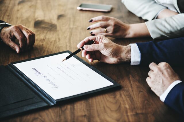 A group of people sits at a wooden table, with one person holding a pen above a marriage certificate. A phone rests nearby, hinting at consulting a divorce attorney. The focus is on the document and the hands, capturing the gravity of this official signing process.