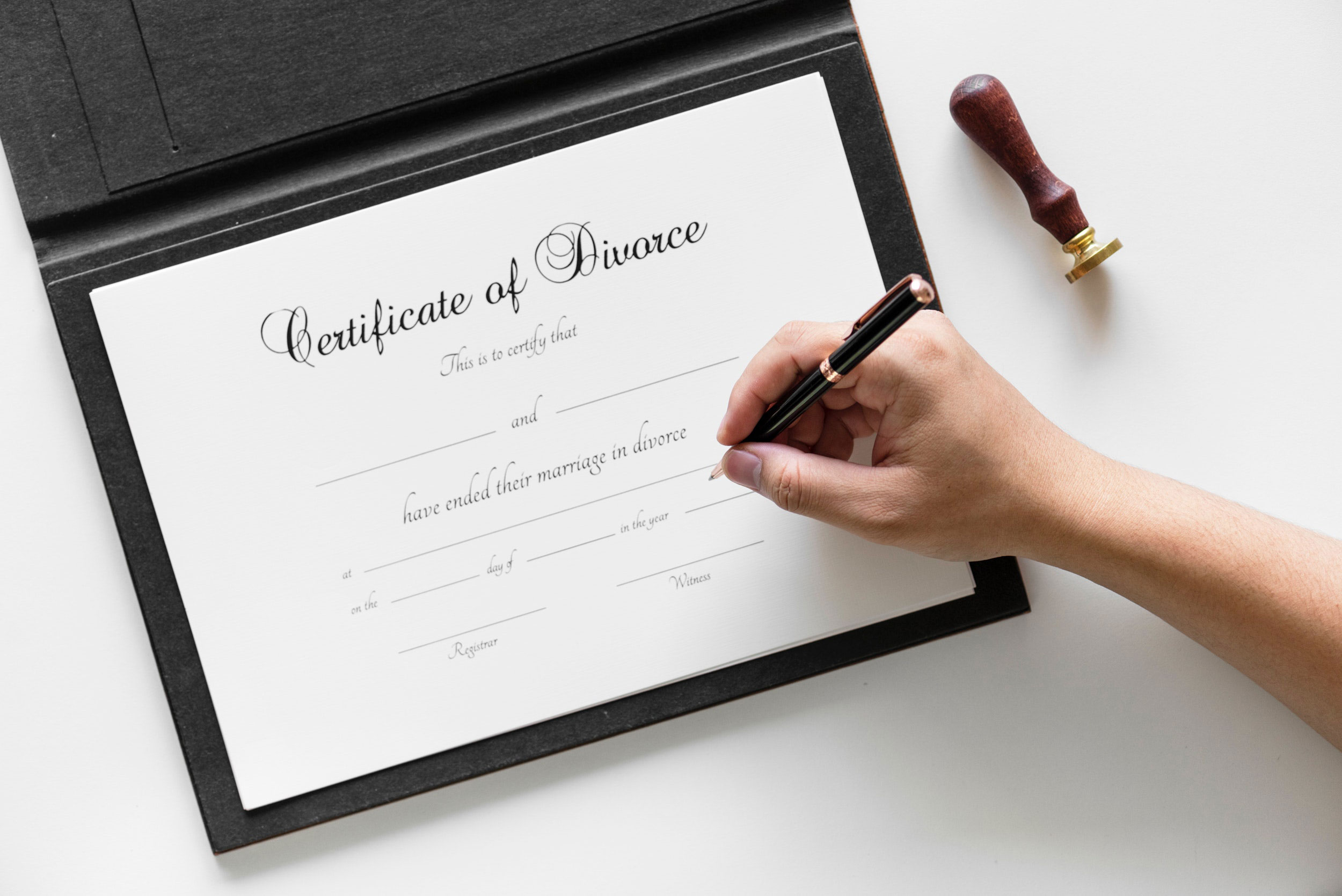 A person is using a pen to fill out a "Certificate of Divorce" on white paper, which a divorce mediator had partially completed. Nearby, a wooden stamp with a brass end rests on the white surface.