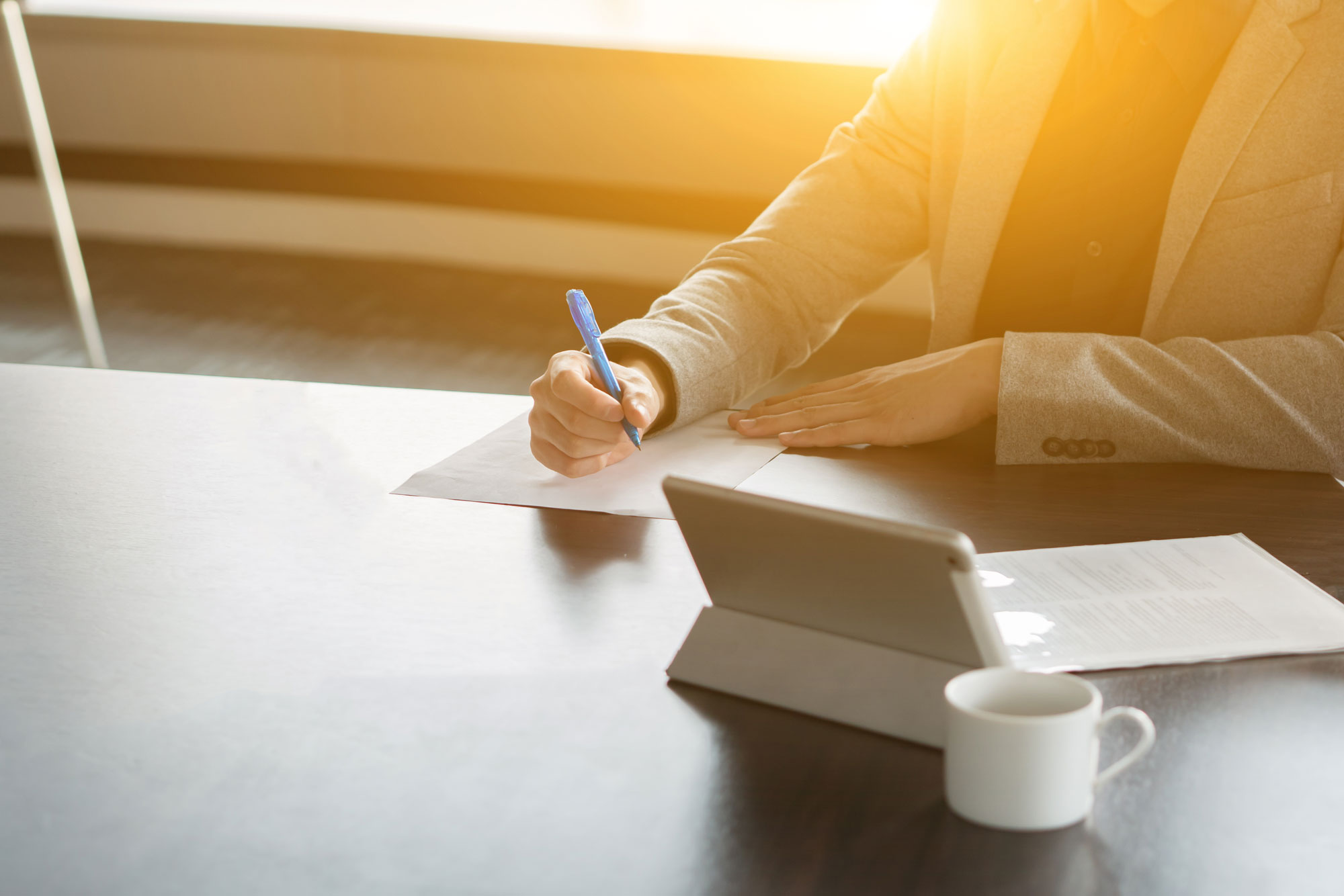 A person in a suit writes on paper at a desk with a tablet and a white coffee cup, resembling the focus and precision of estate planning attorneys. Sunlight streams through the window, casting a warm glow throughout the room.