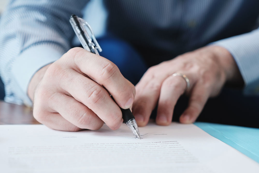A person wearing a blue striped shirt is writing with a black pen on a white sheet of paper. The focus is on the hand and pen, with a blurred background. The person is wearing a ring on their left hand.