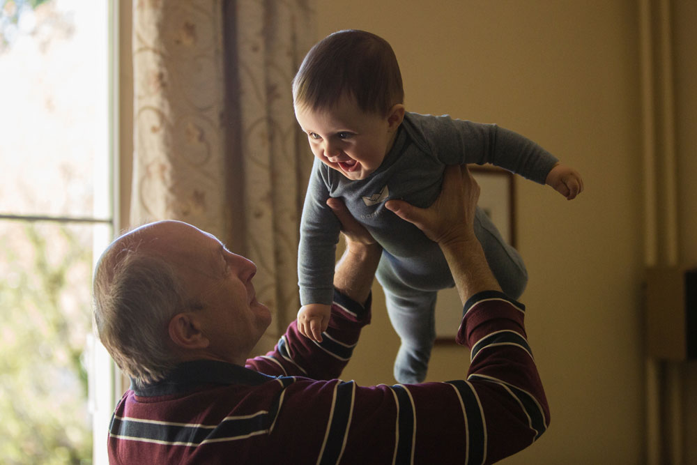 An elderly man wearing a striped sweater joyfully lifts a smiling baby in a blue onesie. The baby looks excited, with arms outstretched. They are indoors near a window with curtains in the background.