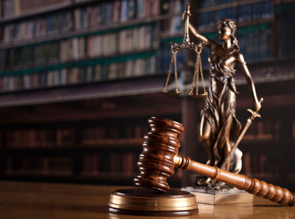 A gavel rests on a wooden sounding block in the foreground. In the background, a statue of Lady Justice holding scales and a sword stands. Shelves filled with books are blurred in the distance, suggesting a legal or courtroom setting.