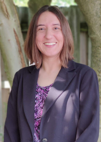 Person with shoulder-length brown hair smiles, standing outdoors in front of a tree. They wear a dark blazer over a purple patterned top. Sunlight filters through the branches, creating dappled light on their outfit.
