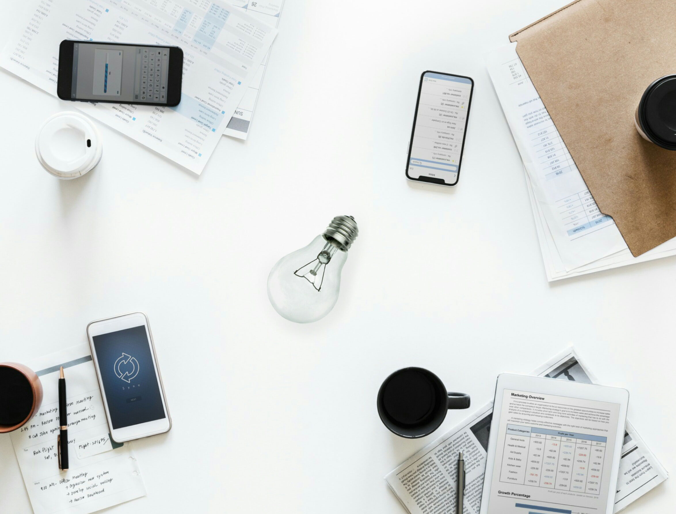 A white desk features a light bulb at the center, surrounded by smartphones, papers, a closed laptop, and coffee cups—creating a productive hub for intellectual property attorneys engaged in collaborative brainstorming.