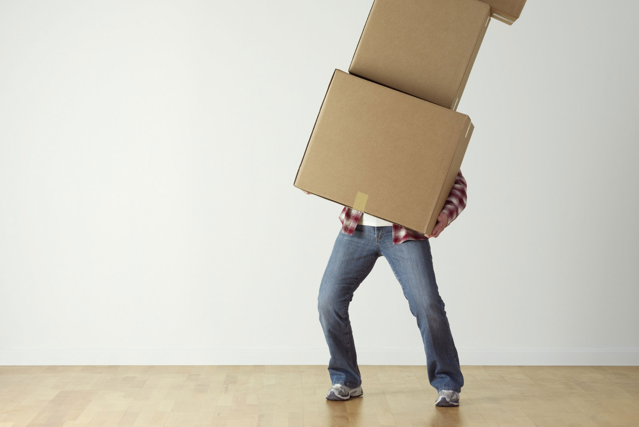 A person in jeans and a plaid shirt struggles to balance while carrying two large cardboard boxes stacked high, obscuring their face. Perhaps they should call a relocation attorney for some logistical advice as they stand on a wooden floor against a plain white wall.