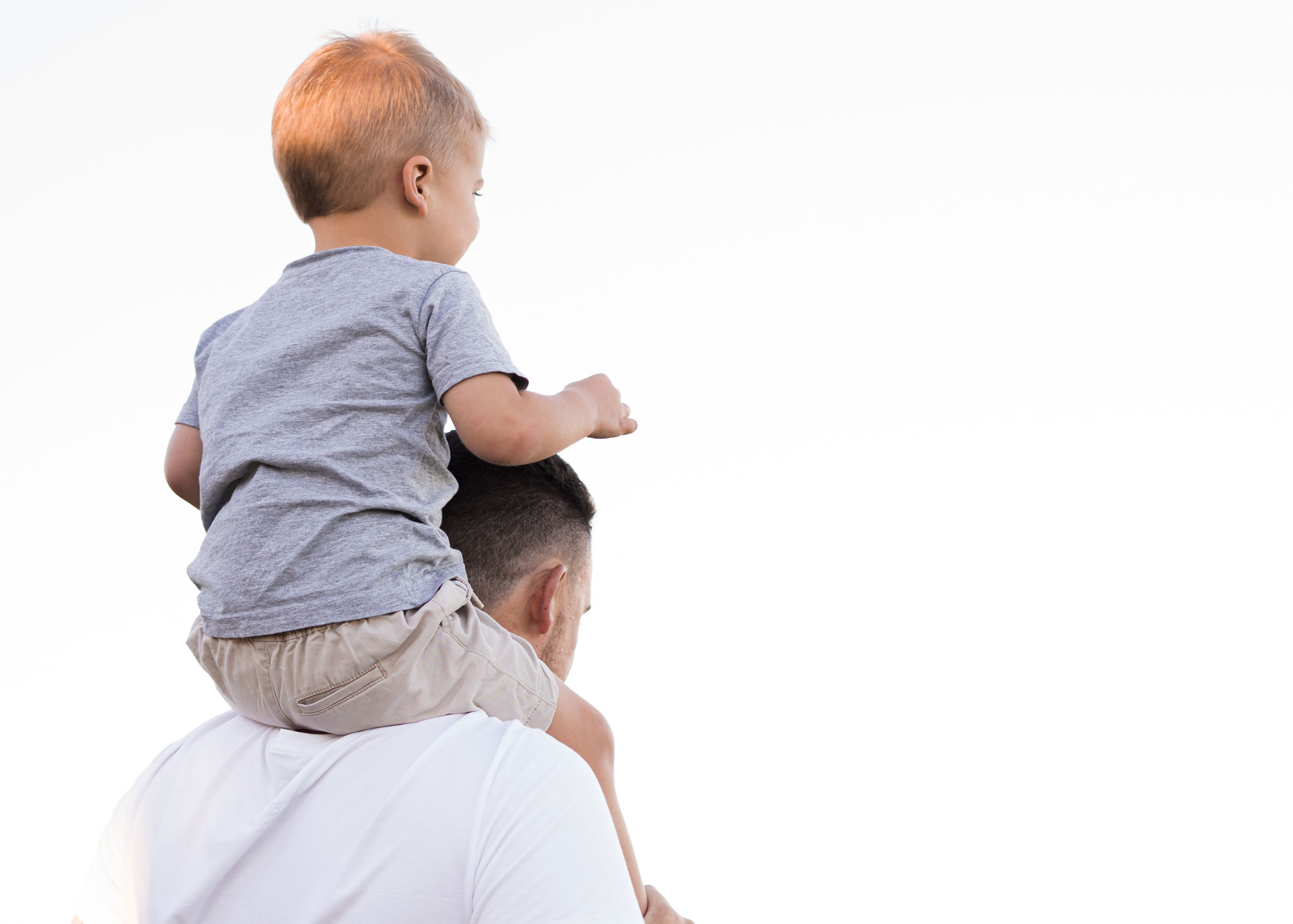 A child with blonde hair sits on an adult's shoulders, enjoying quality parenting time. The child wears a gray shirt and beige shorts, while the adult in a white shirt gazes at the clear blue sky in the background.