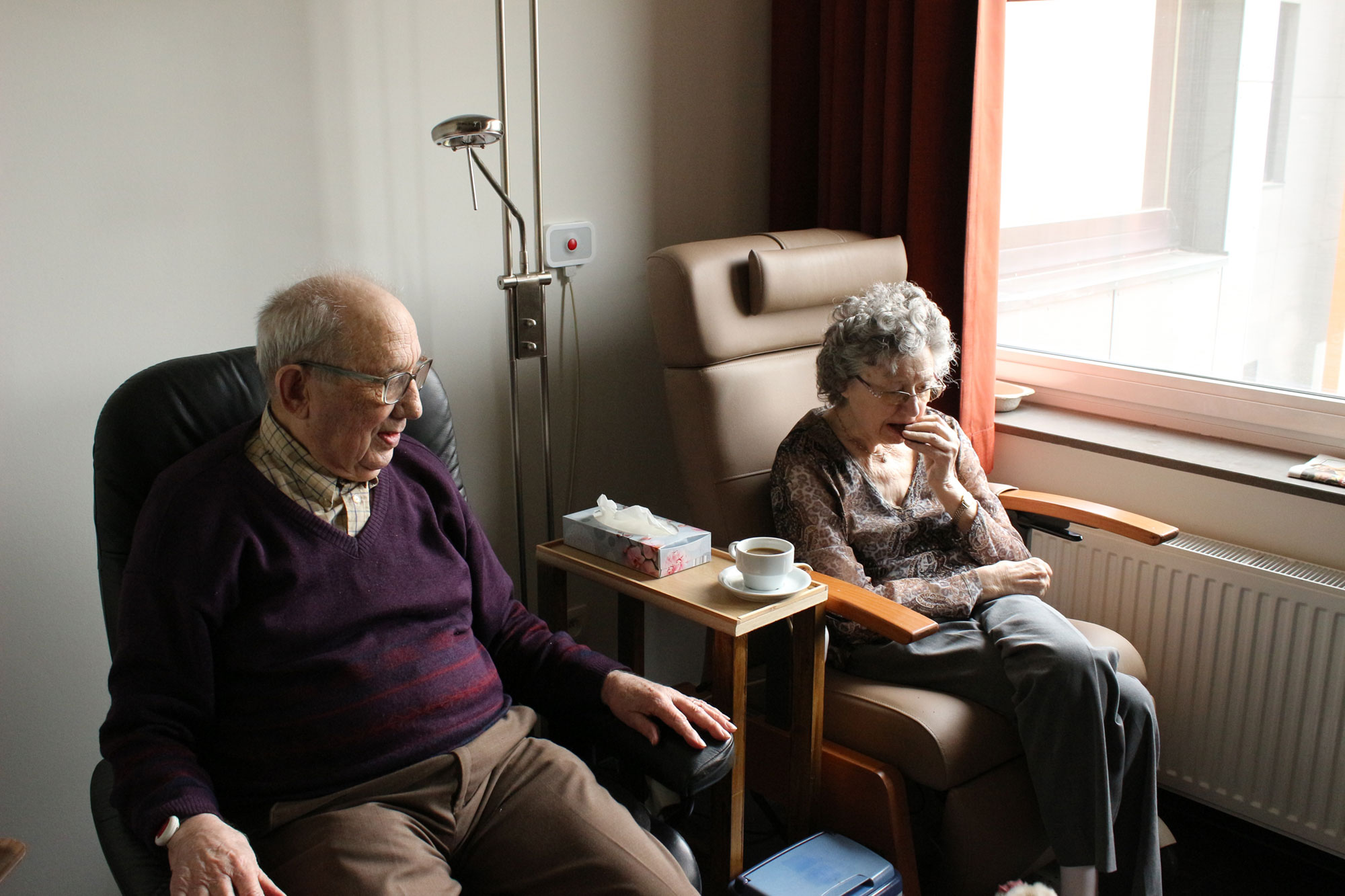 An elderly man and woman sit side by side in armchairs near a window, contemplating life’s decisions. The woman gently touches her mouth, a cup of coffee resting nearby. Close to the man sits a box of tissues, as if considering an important conversation with Power of Attorney lawyers. The room is softly lit.