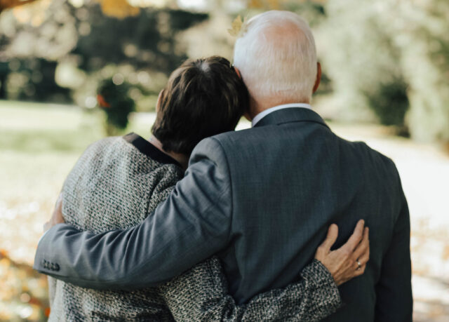 An elderly couple is seen from behind, tenderly embracing each other amidst the serene atmosphere of a park. Dressed in formal attire, with the man in a suit and the woman in a patterned jacket, they exude an air of dignity akin to seasoned probate attorneys navigating life's intricate estate pathways.
