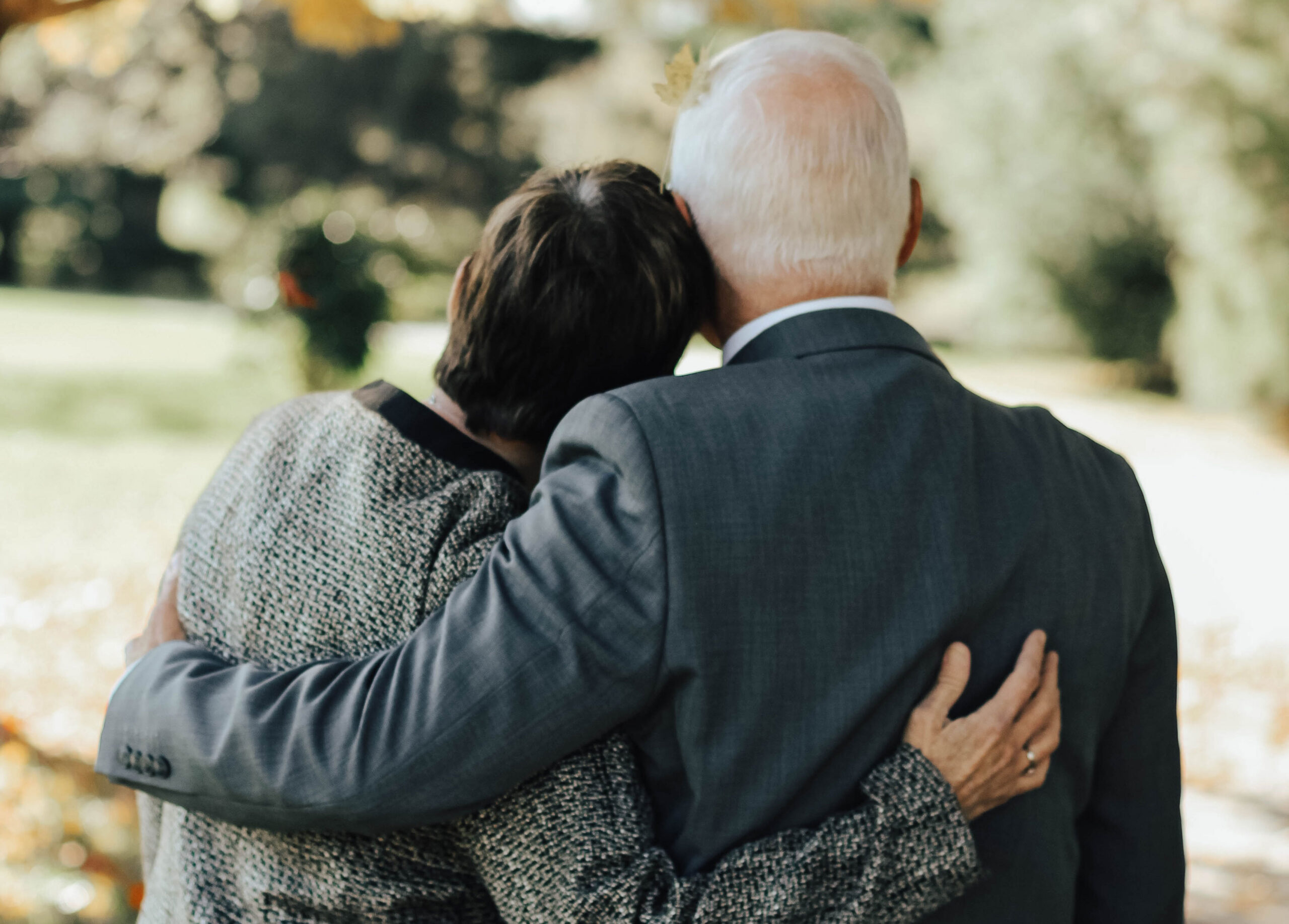 An elderly couple is seen from behind, tenderly embracing each other amidst the serene atmosphere of a park. Dressed in formal attire, with the man in a suit and the woman in a patterned jacket, they exude an air of dignity akin to seasoned probate attorneys navigating life's intricate estate pathways.