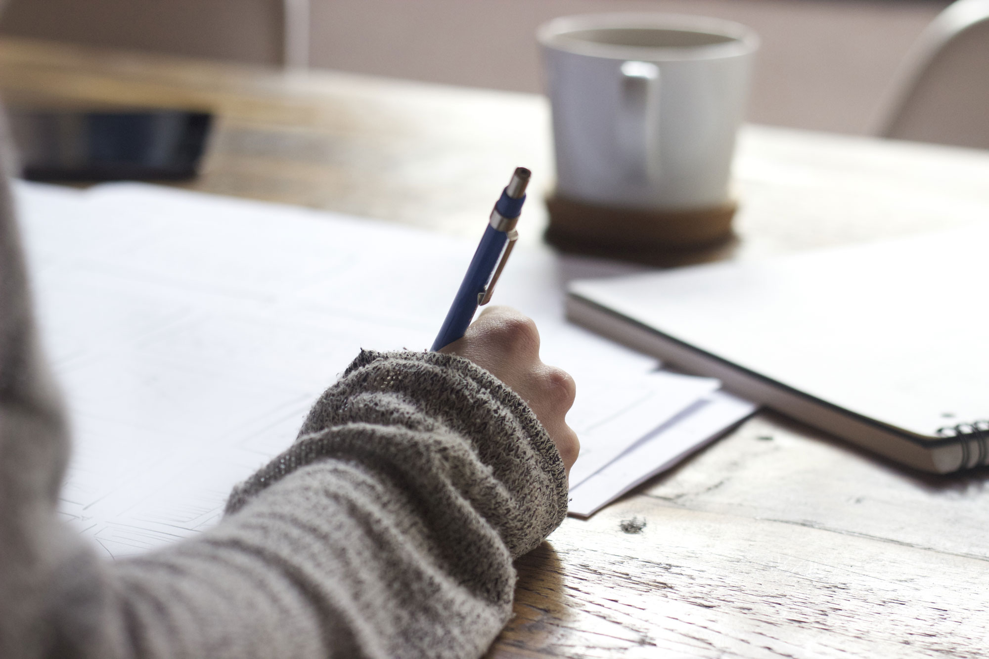 A person in a sweater is writing on paper at a wooden table, perhaps drafting important documents. A mug sits nearby, along with a closed notebook. The scene suggests a calm, focused moment of work—for all we know, they might be preparing as a wills attorney.