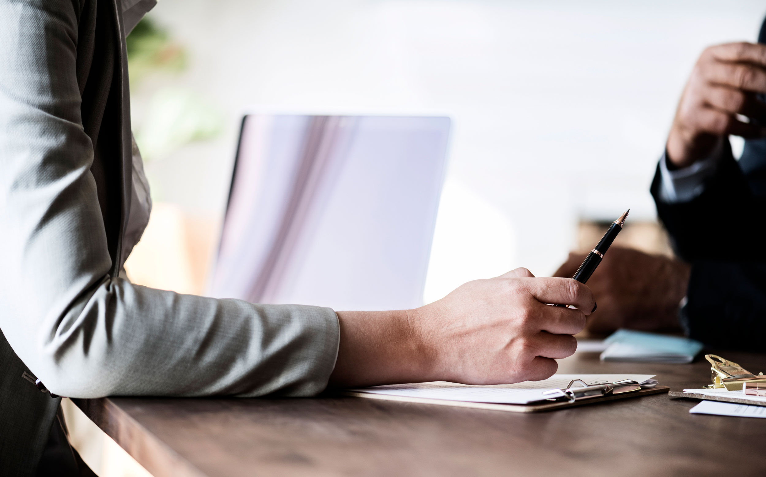 Close-up of two people sitting at a wooden table. One person's hand, possibly a trusts attorney, is holding a pen over a notebook, while a laptop is open in the background. The other person's hand is partially visible, holding a cup. They appear to be deep in discussion or meeting.