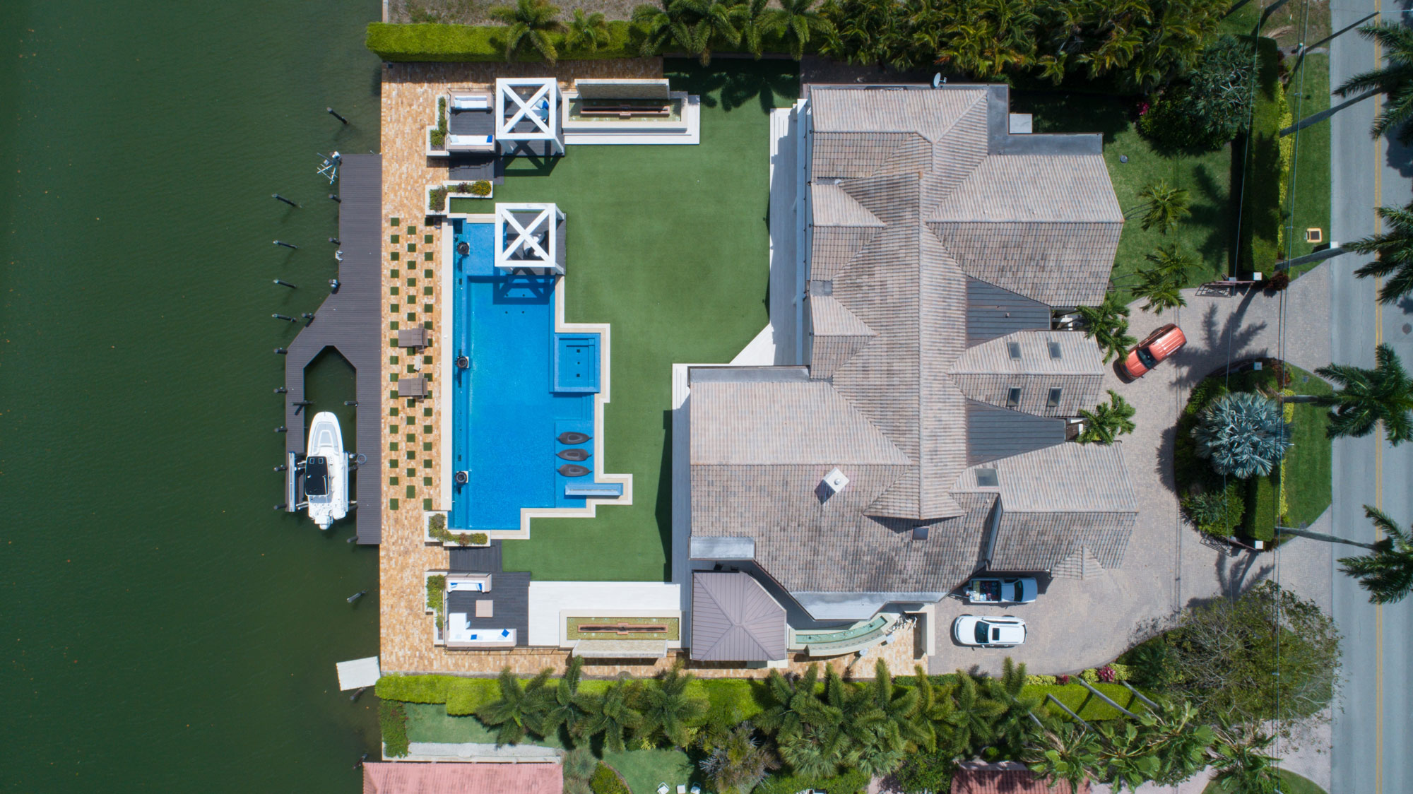 Aerial view of a waterfront estate belonging to a high net worth divorce attorney, featuring a grand house with a tiled roof, rectangular swimming pool, lush green lawn, docked boat surrounded by palm trees, and a driveway lined with parked cars near the entrance.