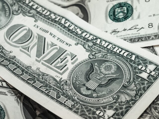 Close-up of multiple U.S. one-dollar bills overlapping each other. The prominent bill features "ONE DOLLAR" and the Great Seal of the United States with the eagle emblem visible. The words "IN GOD WE TRUST" are also displayed.