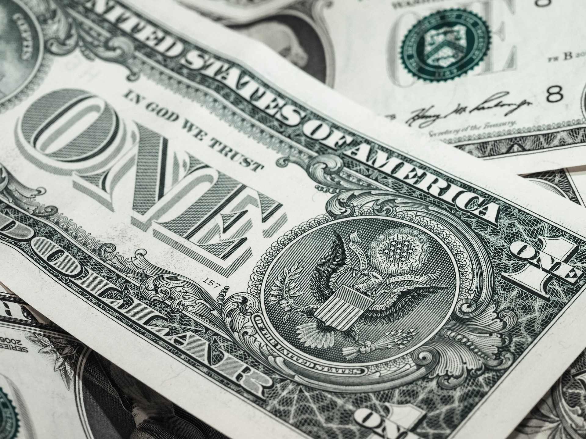 Close-up of multiple U.S. one-dollar bills overlapping each other. The prominent bill features "ONE DOLLAR" and the Great Seal of the United States with the eagle emblem visible. The words "IN GOD WE TRUST" are also displayed.