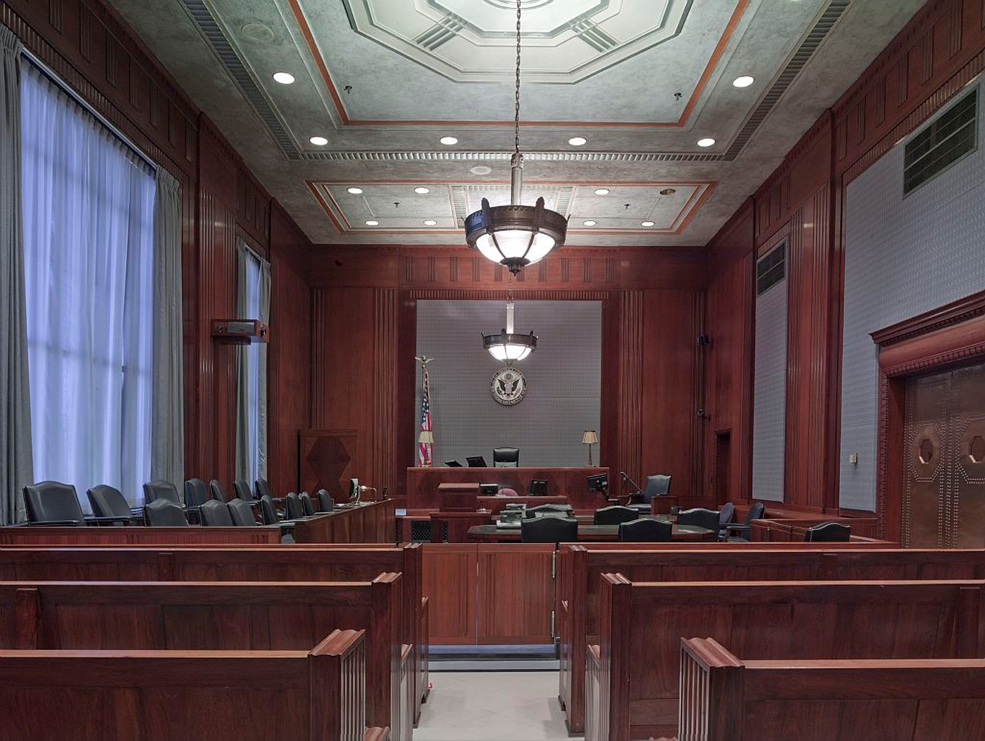 A courtroom with wooden furnishings, empty seats for the jury, and a judge's bench. American flags flank the central podium. Overhead, chandeliers illuminate the room, and tall curtains cover large windows.