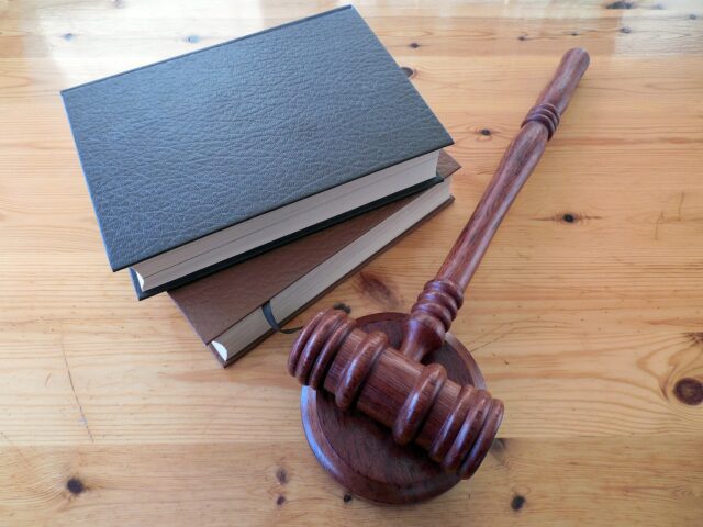 A wooden gavel rests on a matching sound block on a wooden table. Three hardcover books with plain covers are stacked nearby. The scene suggests a legal or judicial setting.