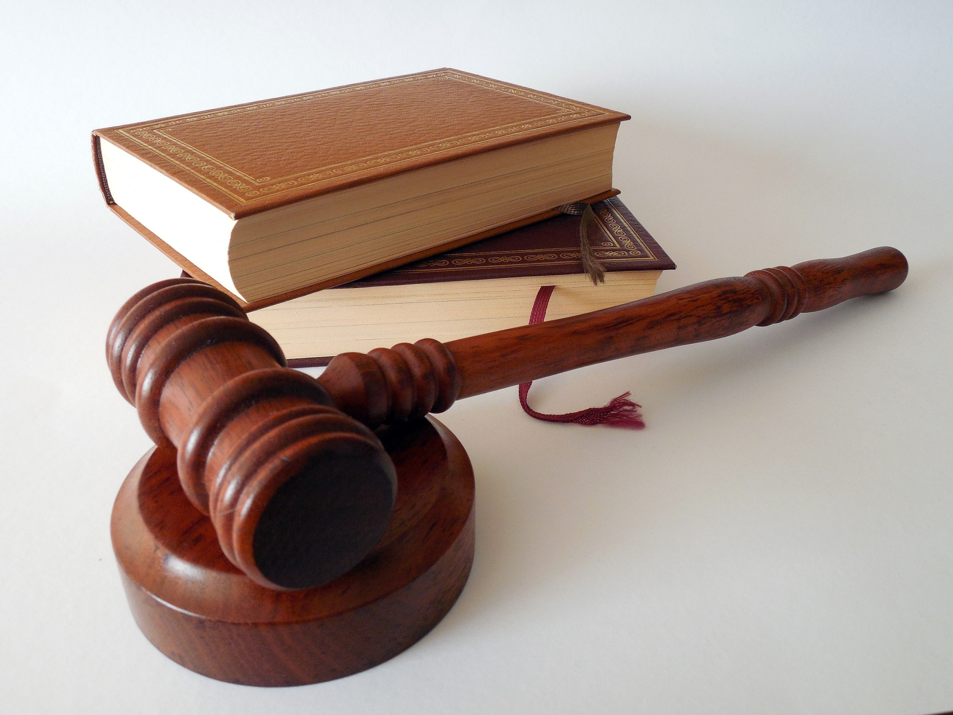 A wooden gavel rests on a matching sound block, positioned in front of two closed, brown hardcover law books. The background is plain and white, highlighting the legal theme of the objects.