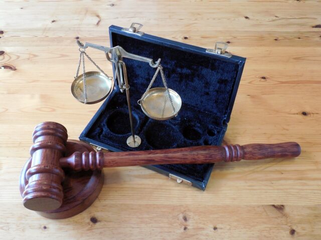 A wooden gavel rests on a sound block next to a set of brass balance scales. The scales are placed in a black velvet-lined case on a wooden surface.