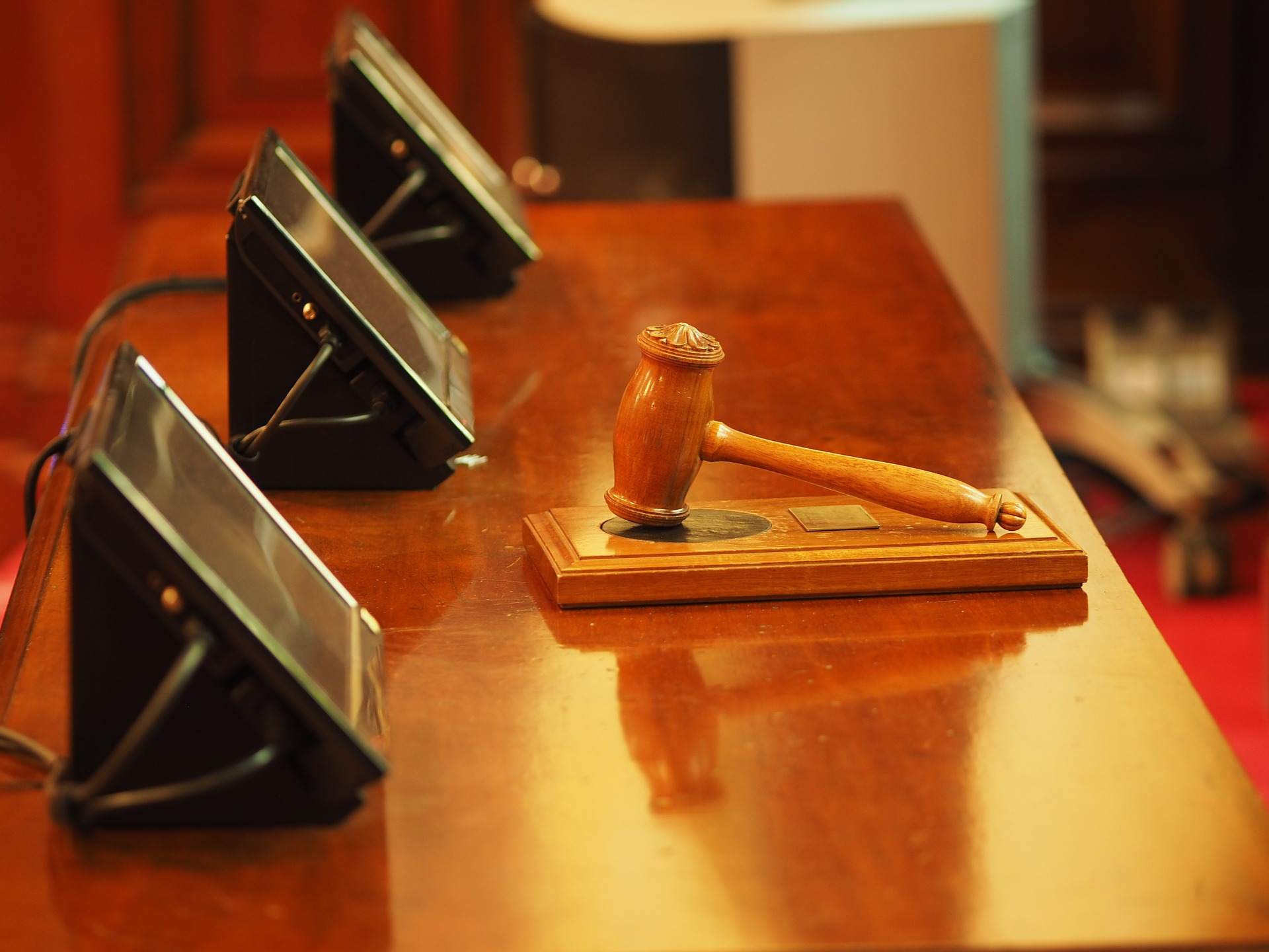 A wooden gavel rests on a sound block atop a polished wooden table. Three black electronic devices with screens are positioned nearby, angled towards the gavel. The background shows part of a wooden panel and red carpet.