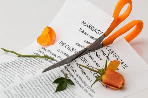A pair of orange-handled scissors rests on a torn marriage certificate, accompanied by a partially cut orange rose. The scene suggests themes of separation or divorce.