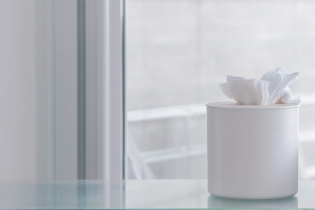 A white tissue box with tissues protruding sits on a glass surface. The background is softly blurred, featuring a large window, creating a minimalistic and clean aesthetic.