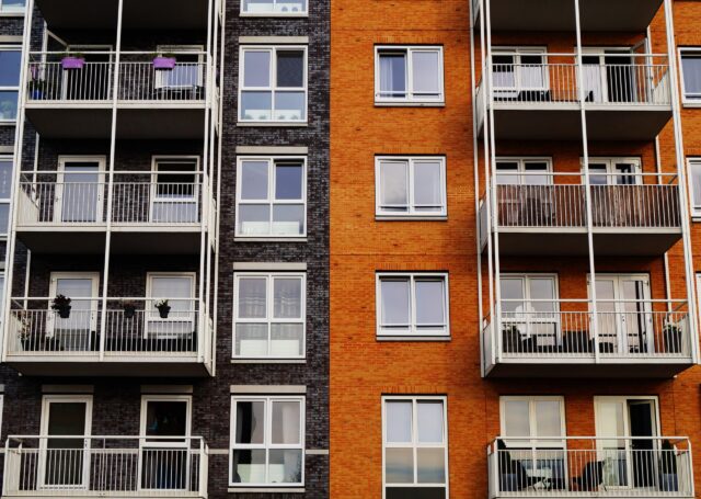 A view of a modern apartment building with two contrasting façades. The left side features dark brick with balconies displaying plants. The right side is orange brick with similar balconies, creating a symmetrical architectural design.