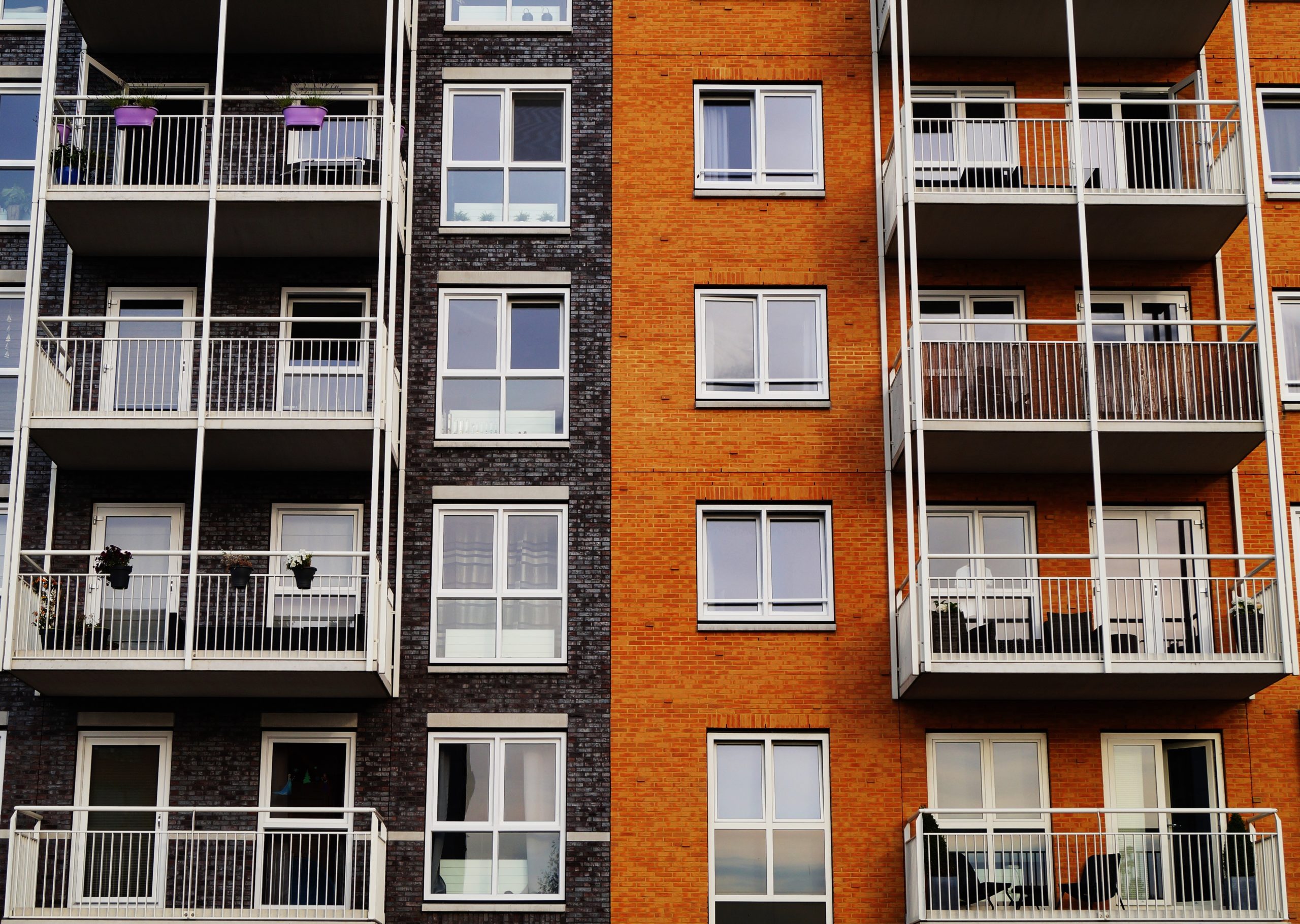 A view of a modern apartment building with two contrasting façades. The left side features dark brick with balconies displaying plants. The right side is orange brick with similar balconies, creating a symmetrical architectural design.