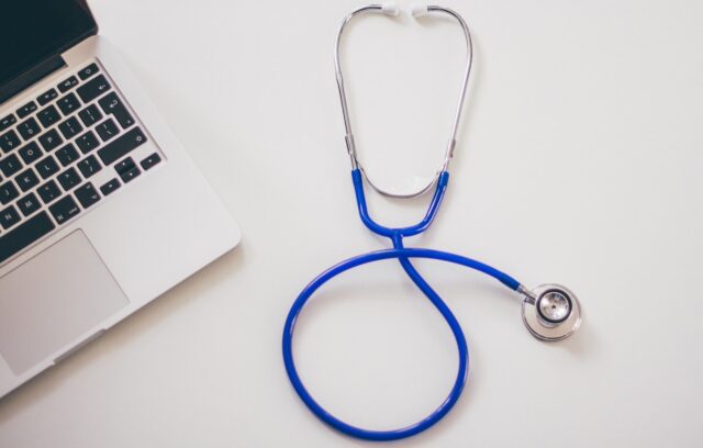 A stethoscope with a blue tube rests on a white surface beside the corner of an open laptop, evoking a medical setting. Nearby, an advance healthcare directive hints at thoughtful planning in patient care.