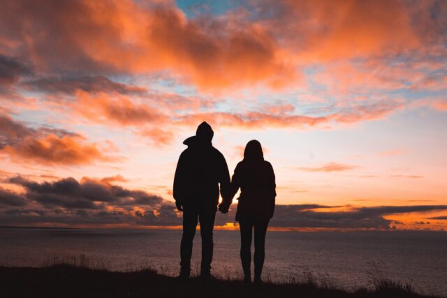 An unmarried couple, silhouetted against a dramatic sunset over the ocean, holds hands as vibrant orange, pink, and purple clouds paint the sky. The scene creates a serene and romantic atmosphere.