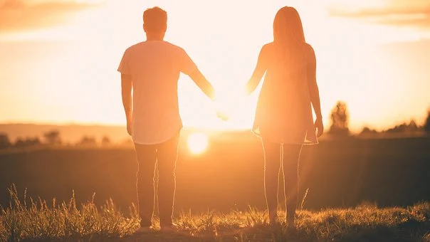 A man and woman stand hand in hand, silhouetted against a bright orange sunset. They are on a grassy field, contemplating estate planning for unmarried couples, facing the horizon with the sun low in the sky, creating a warm and peaceful atmosphere.
