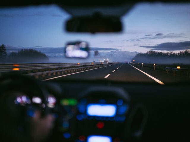 A person with past OWI convictions drives a car on a highway at dusk. The dashboard is slightly blurred, and the road ahead is lined with trees, illuminated by streetlights. The sky is darkening with a few clouds visible, reflecting their desire for a fresh start.