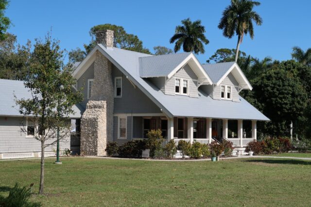 A large gray house with a stone chimney, white trim, and a spacious porch sits on a well-kept lawn perfect for children to play. Small trees dot the grass, while palms and other trees stand in the background beneath a clear blue sky.