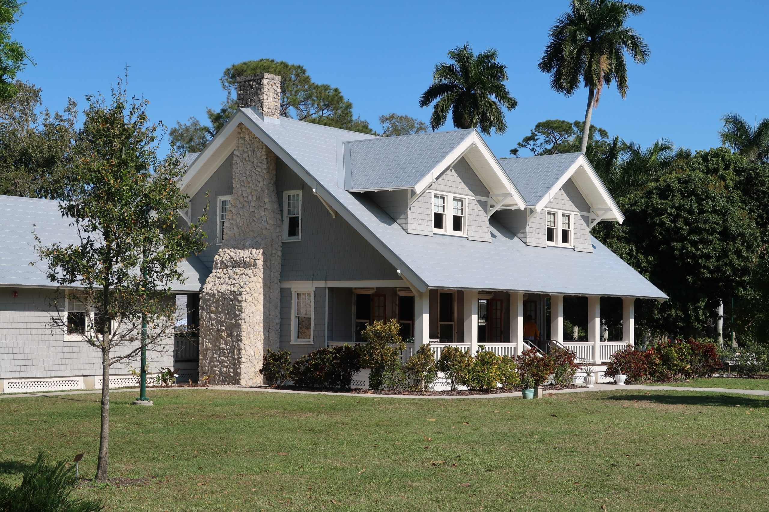 A large gray house with a stone chimney, white trim, and a spacious porch sits on a well-kept lawn perfect for children to play. Small trees dot the grass, while palms and other trees stand in the background beneath a clear blue sky.