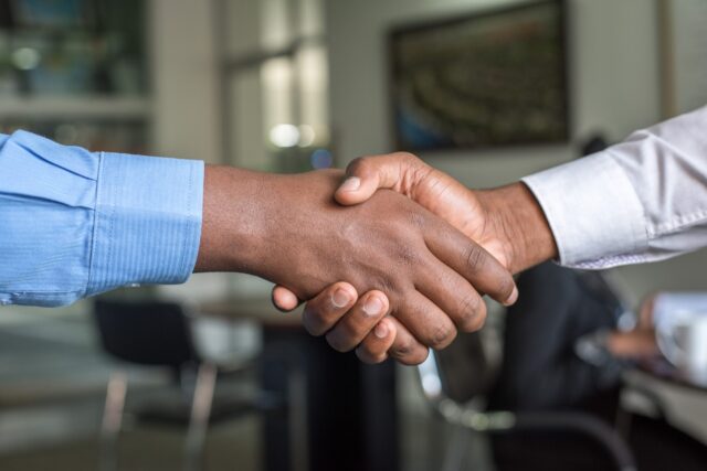 Two individuals engaged in an initial consultation warmly shake hands in an office setting, both clad in long-sleeved shirts—one in blue, the other in white. The blurred background, featuring office furniture, underscores a professional or business environment.